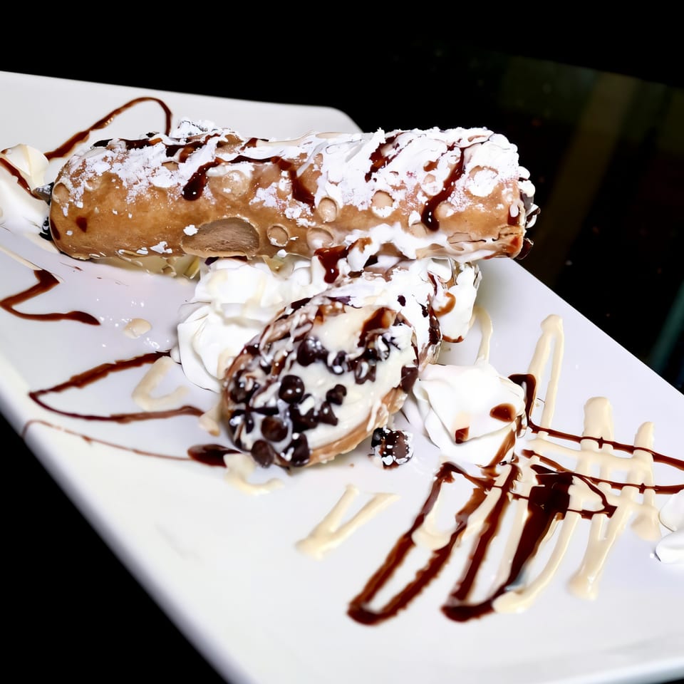 Two cannolis, one large and one small, drizzled with chocolate and powdered sugar on a white plate.