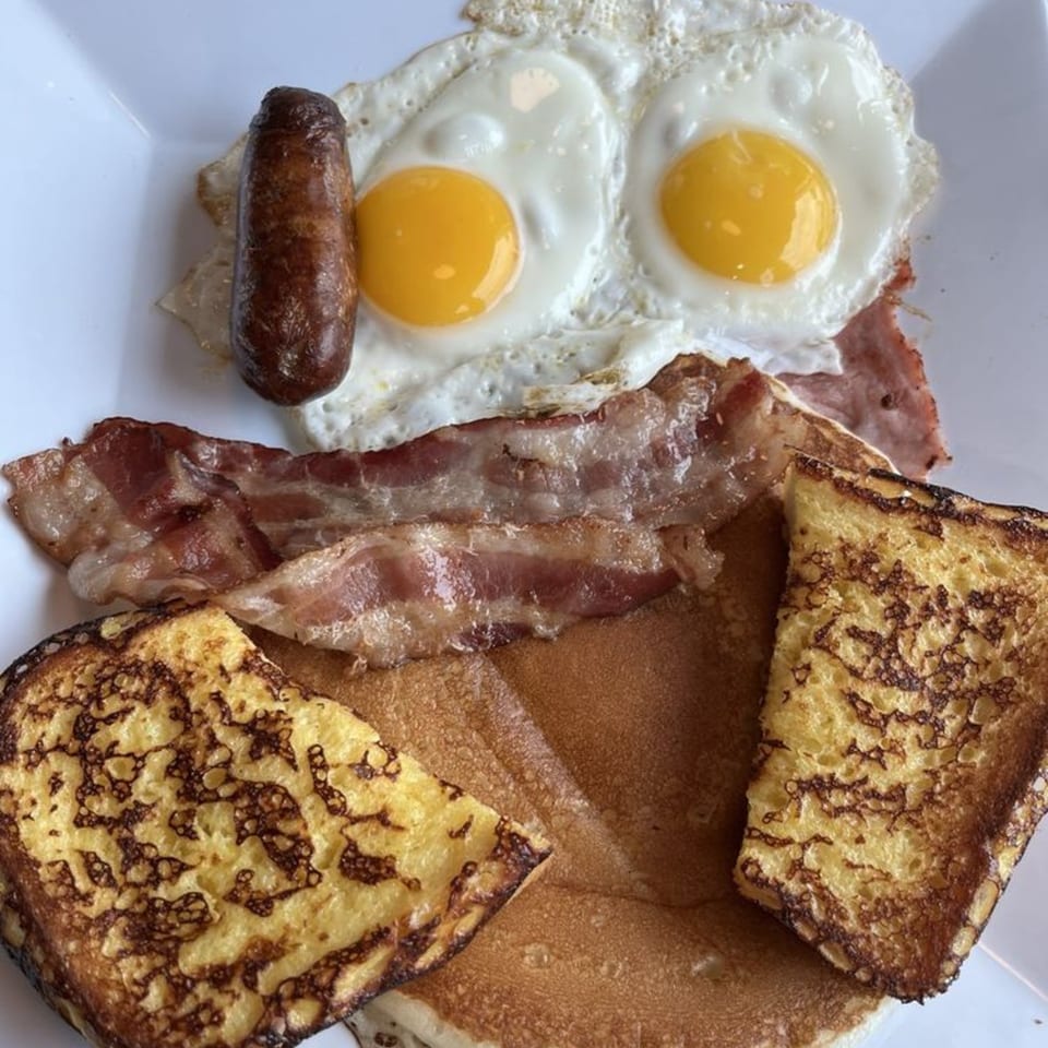 A diner breakfast with sausage, two fried eggs, bacon, French toast, and pancakes on a white plate.