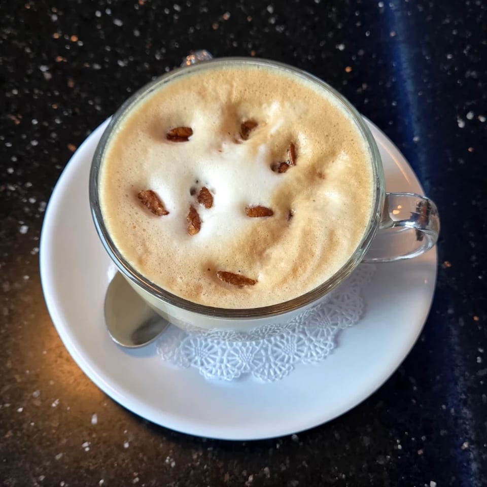 A clear glass mug of frothy coffee with small brown toppings, on a white saucer with a doily.